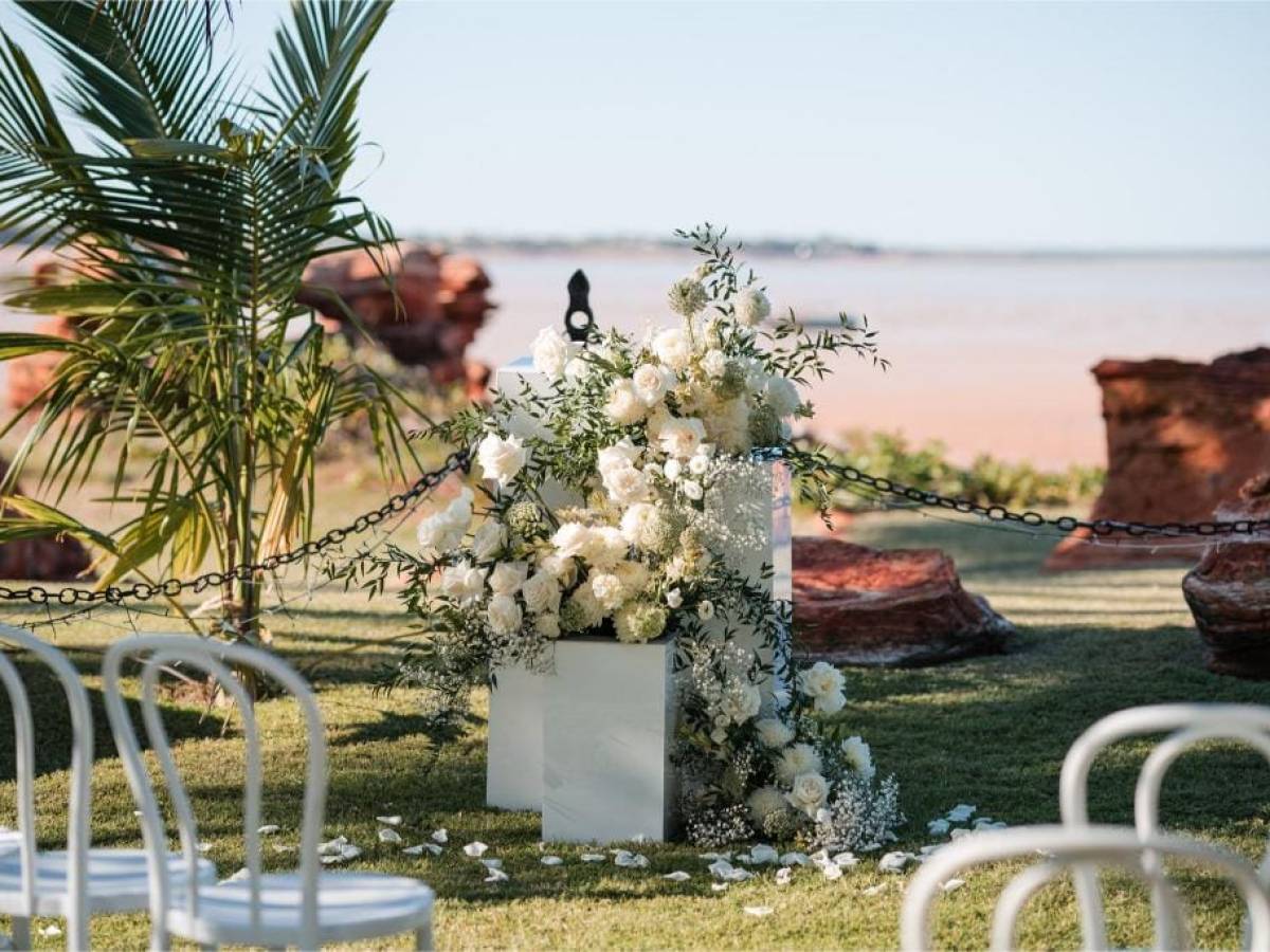 Outdoor wedding setup with white flowers, chairs, and red rocks near a beach.