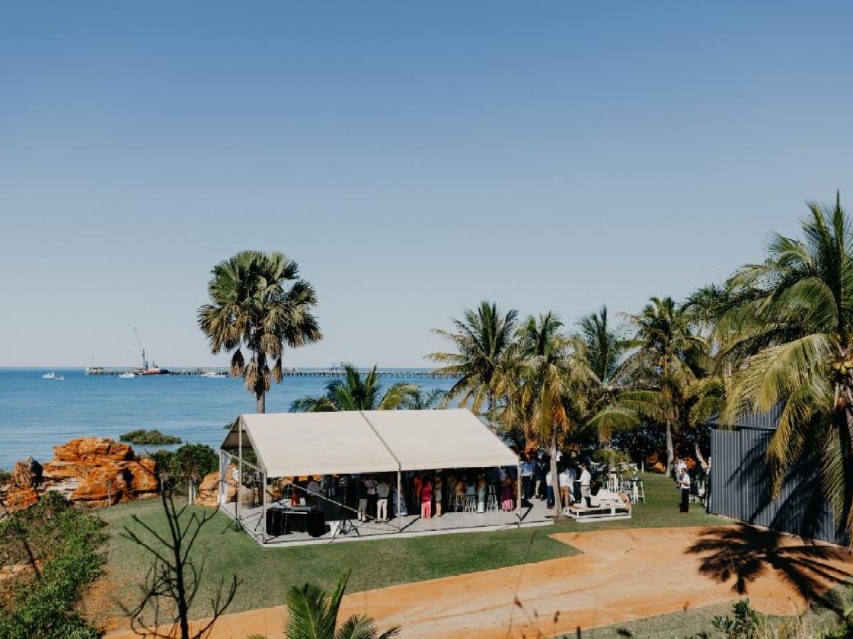 Outdoor event under a tent near palm trees and ocean, with a pier in the background.