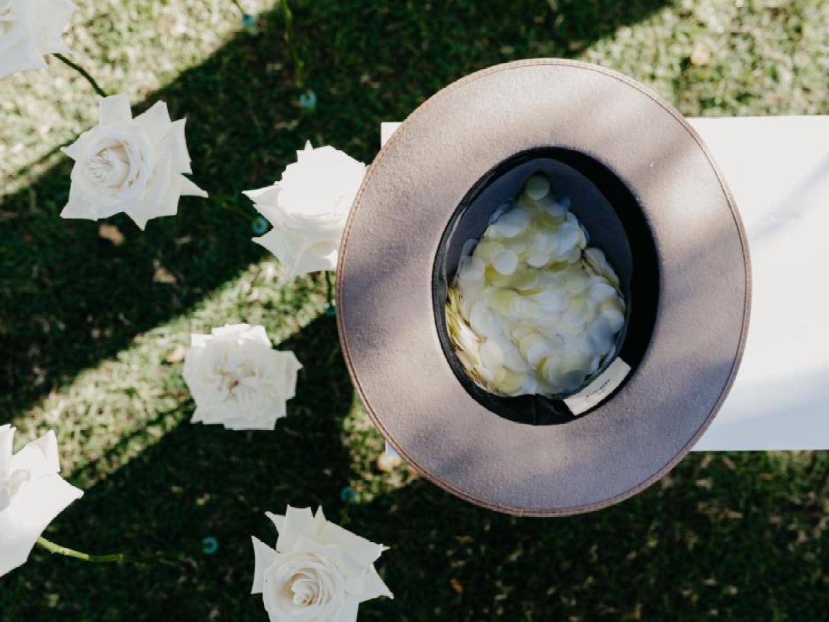 Top view of a brown hat filled with white petals on a table, surrounded by white roses on grass.