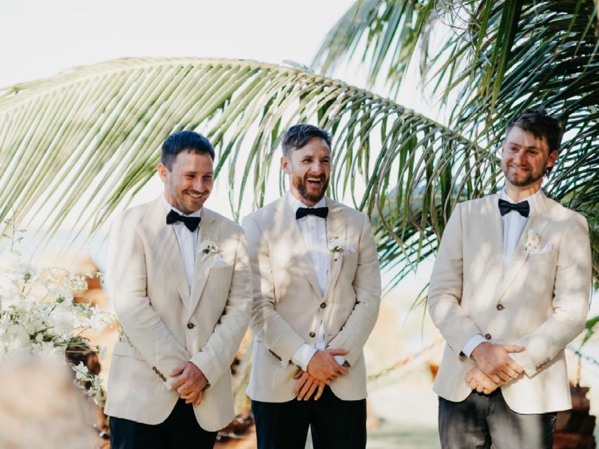Three men in light suits with bow ties smiling under palm leaves at an outdoor event.