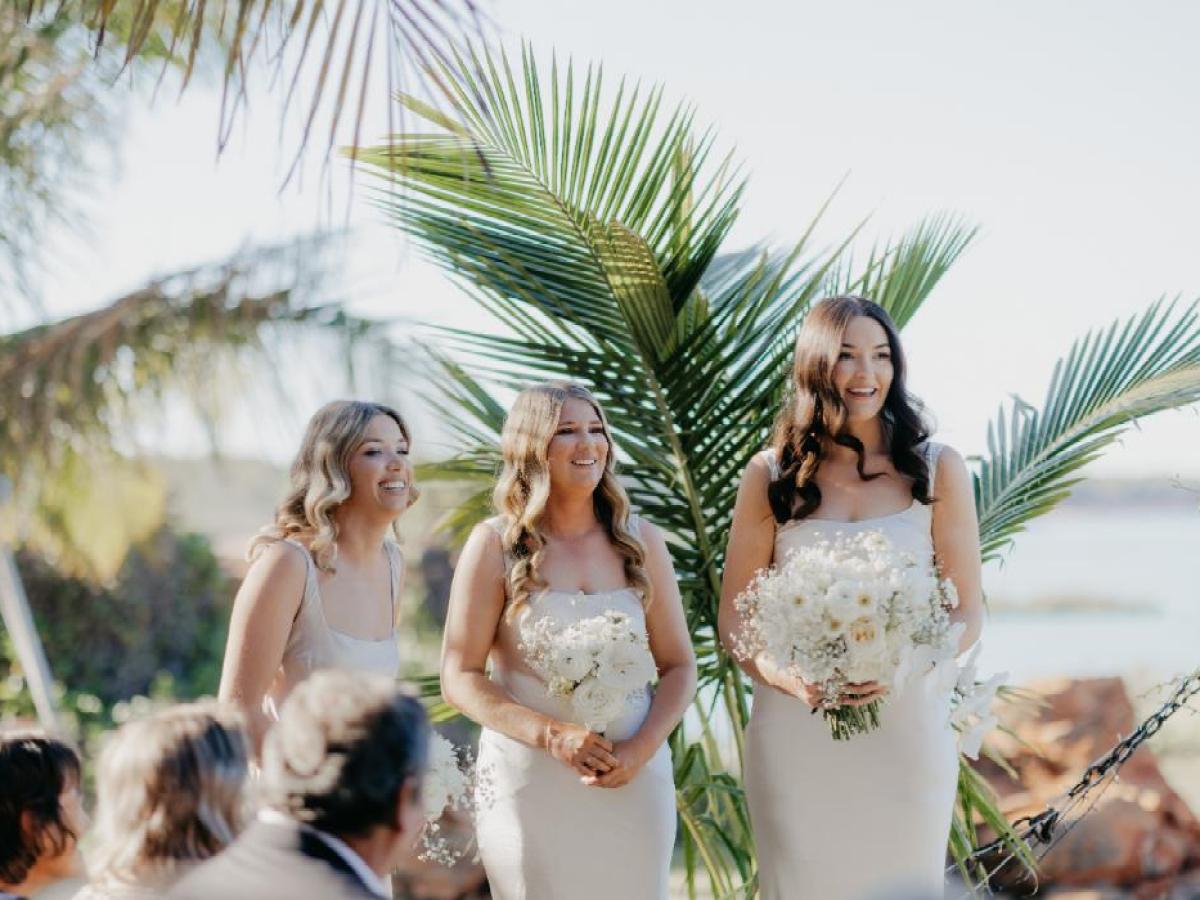 Three women in matching dresses holding flowers at an outdoor event with palm leaves in the background.