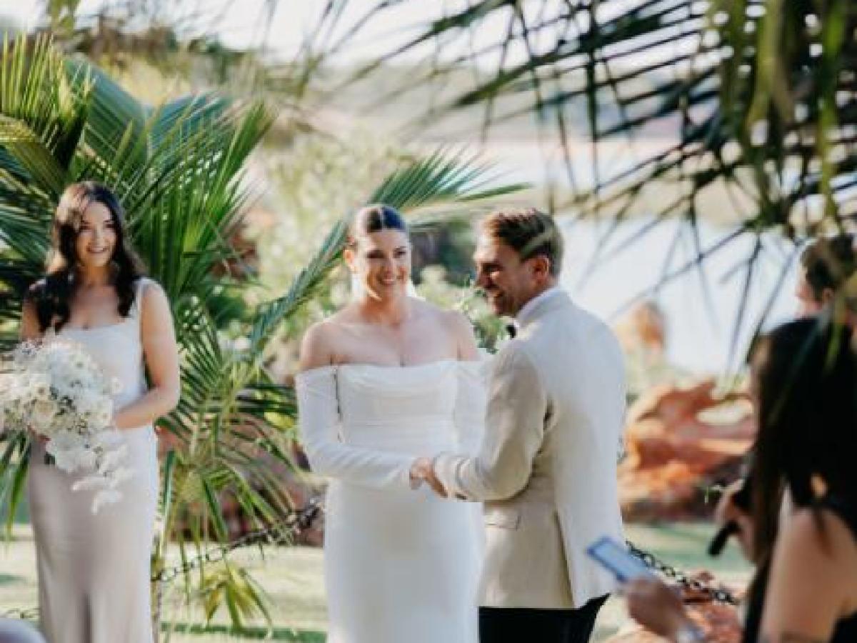 Bride and groom exchange vows outdoors under palm trees with guests seated nearby.