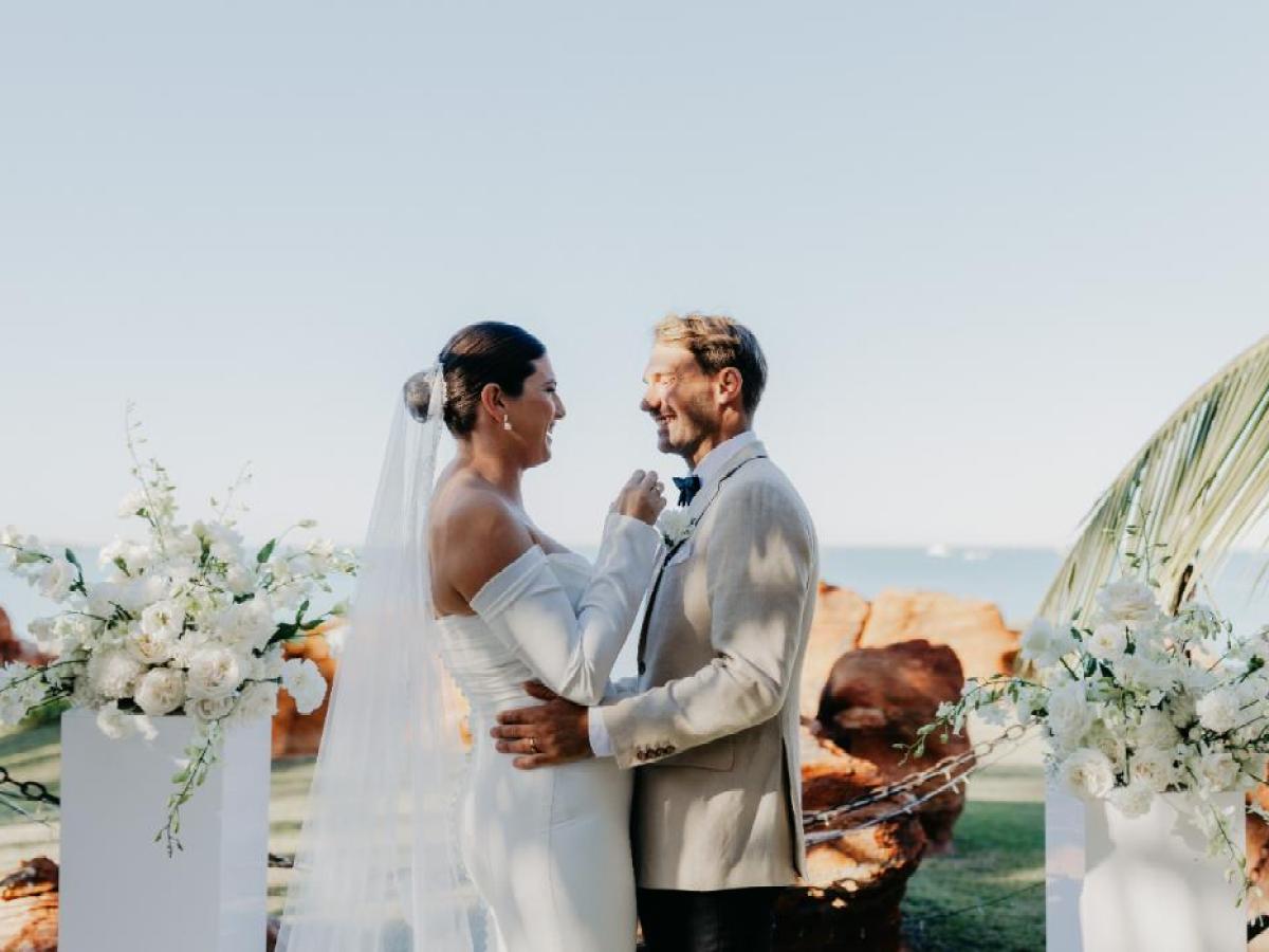 Bride and groom smiling at each other, holding hands, surrounded by white flowers at a beachside wedding.