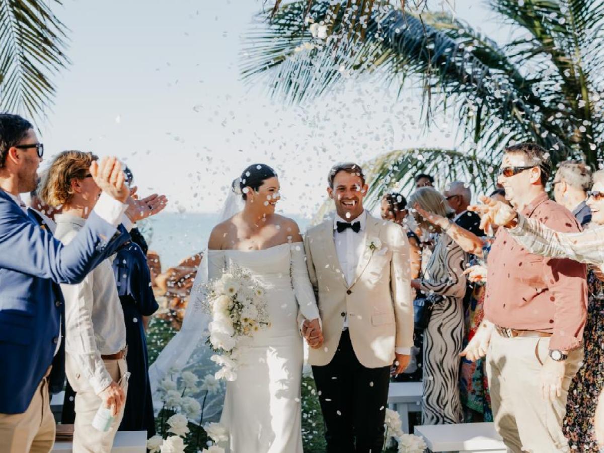 Bride and groom smiling while guests throw confetti at a beach wedding ceremony under palm trees.