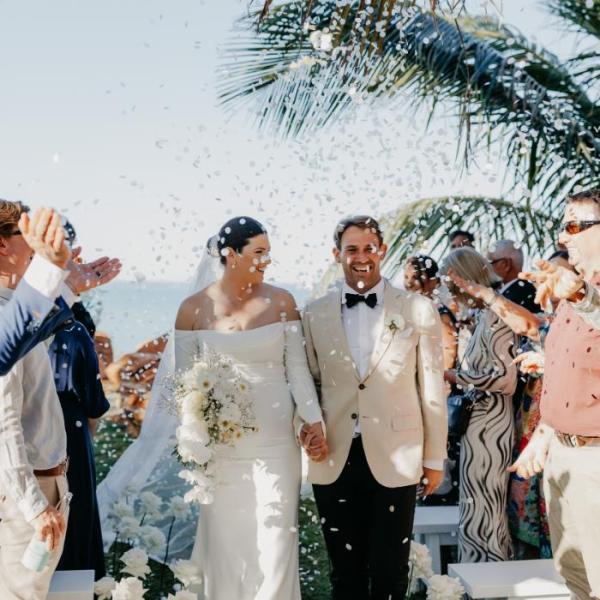 Bride and groom smiling while guests throw confetti at a beach wedding ceremony under palm trees.