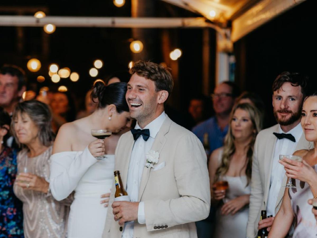Group of people in formal attire, smiling and holding drinks at a celebration event, with string lights overhead.