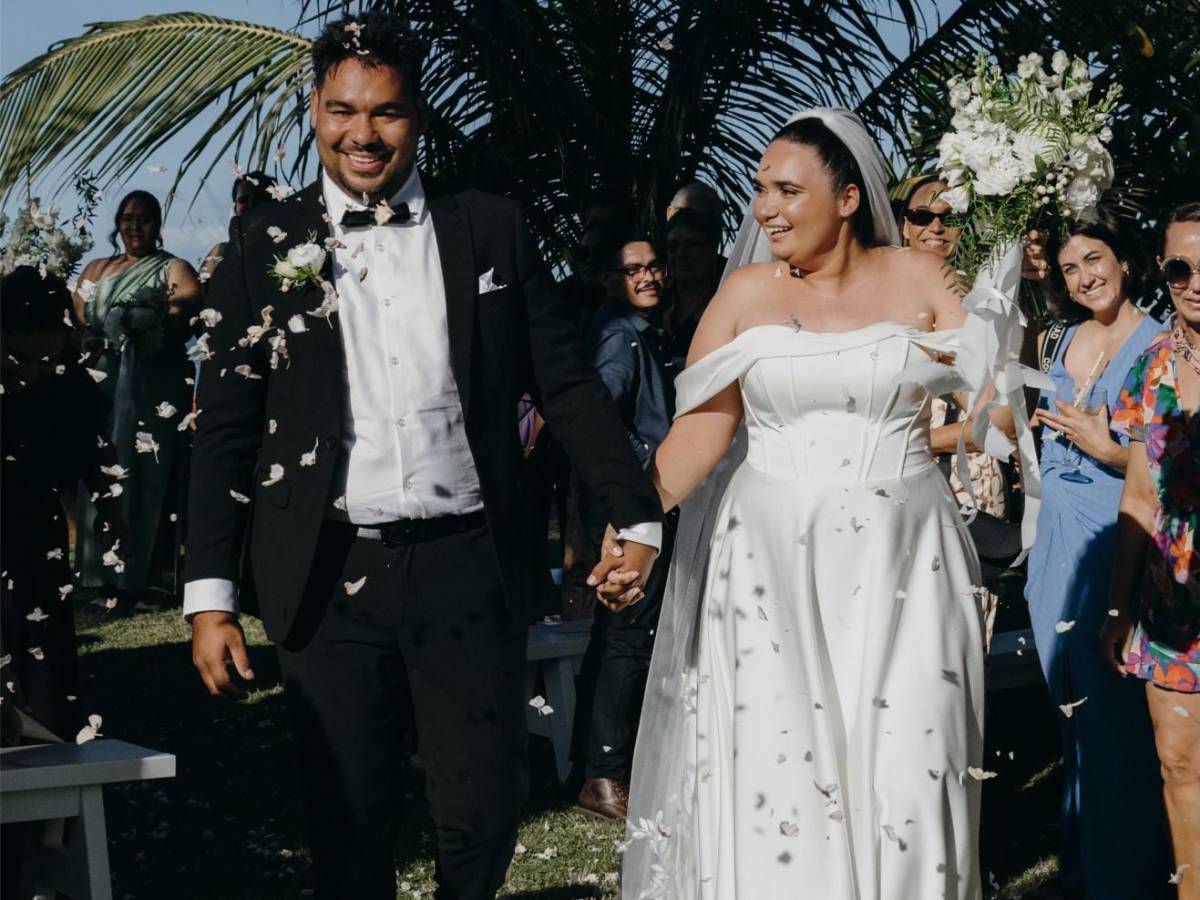 Bride and groom walking under palm trees with guests and falling confetti.
