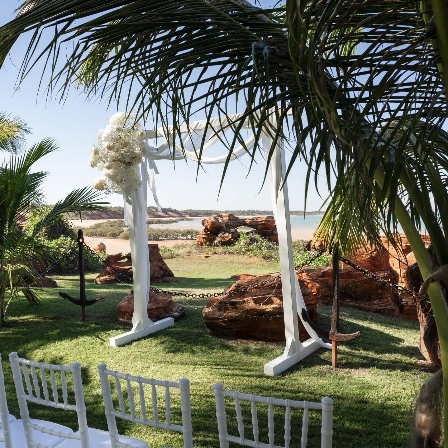 Outdoor wedding arch and chairs on grass with palm trees and ocean view.