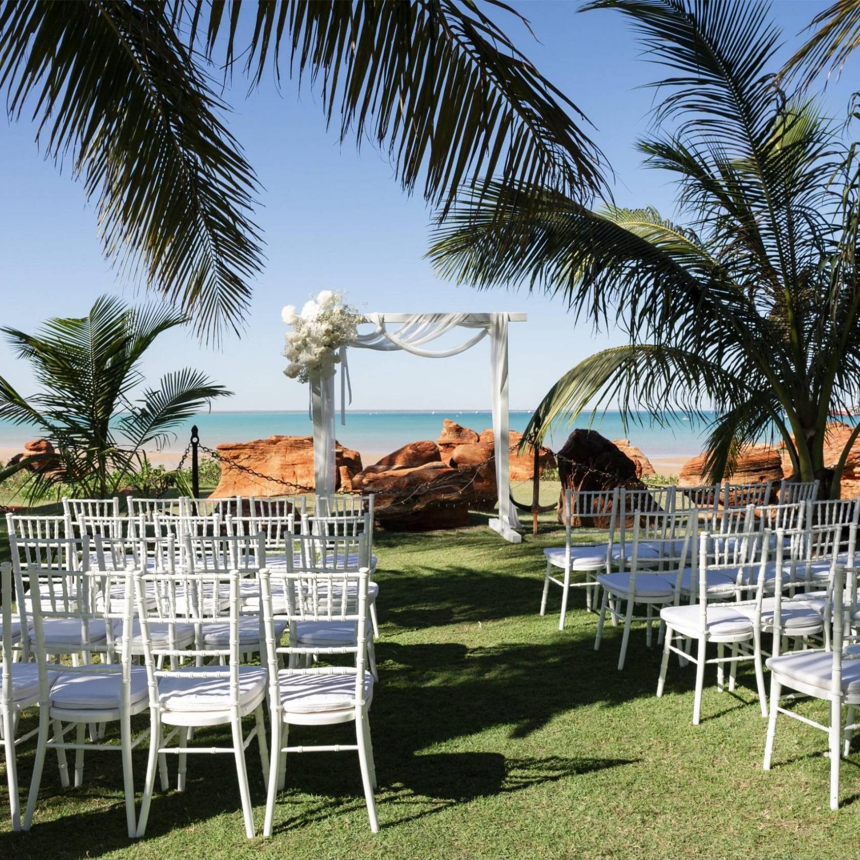 Outdoor wedding setup with white chairs, arch, palm trees, and ocean view.