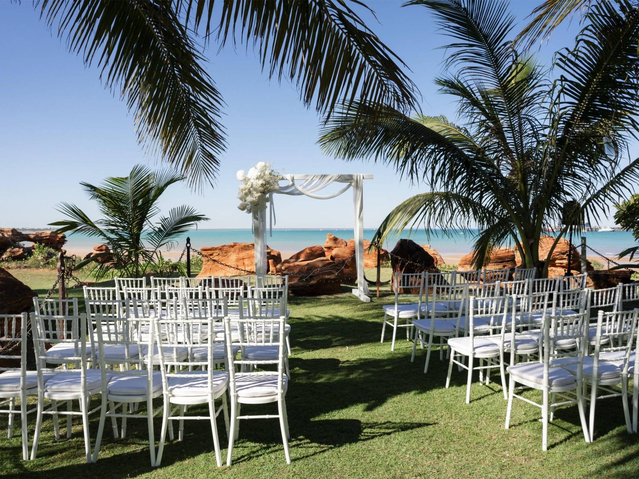 Outdoor wedding setup with white chairs, arch, palm trees, and ocean view.