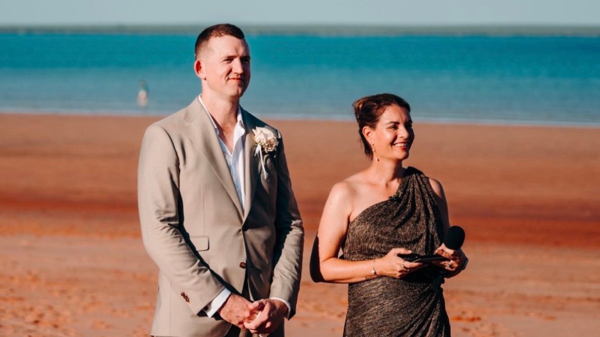 A man and woman stand on a beach, the man in a suit and the woman in a dress.