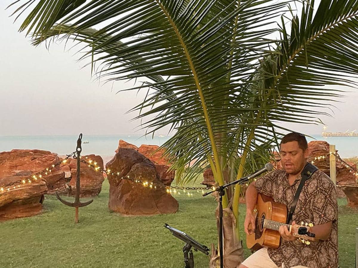 Man playing guitar and singing under palm tree near beach with rocks and string lights.