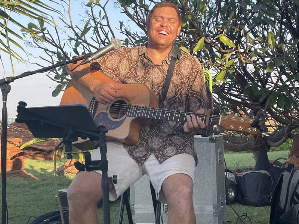 Person playing acoustic guitar outdoors, smiling, with trees and microphone nearby.
