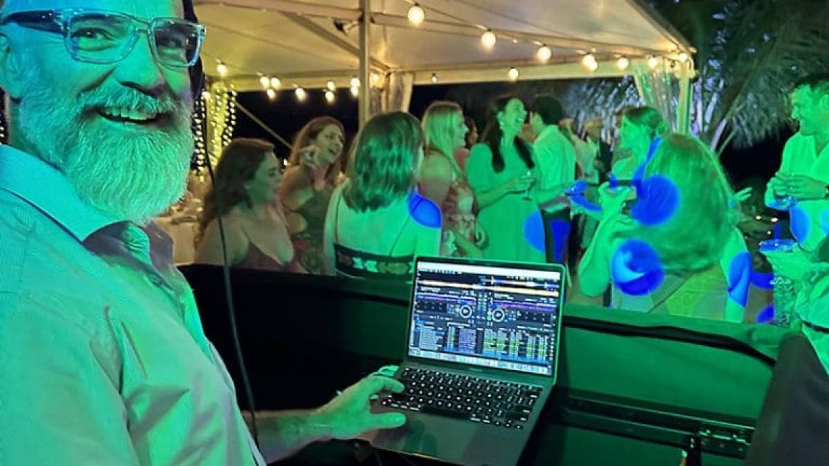 DJ in a tropical setting with laptop and mixer, smiling at a lively party under a tent with string lights.