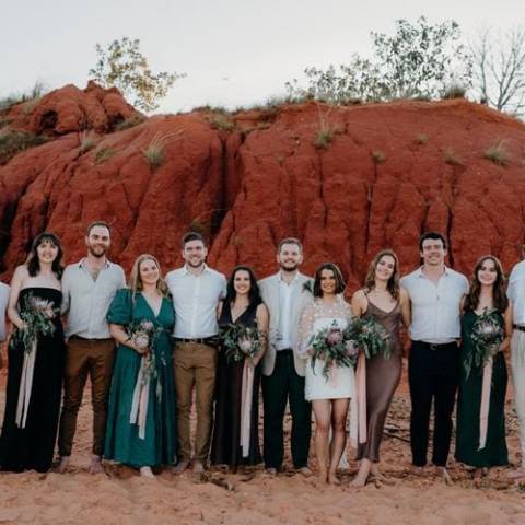 A group poses barefoot in front of red rocks, holding bouquets, with a clear sky above.