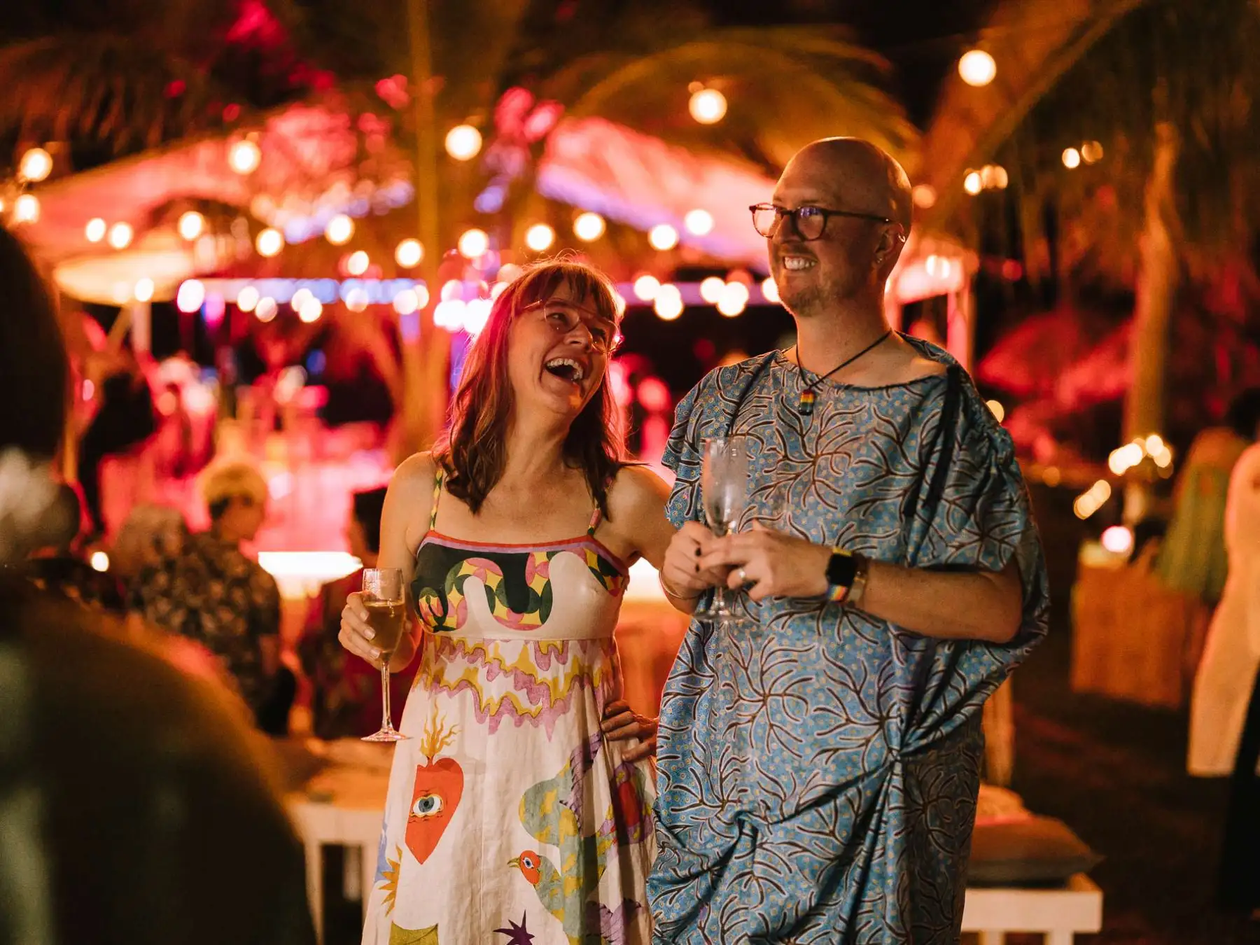 A couple laughing at a festive outdoor night event with colorful lights and people in the background.