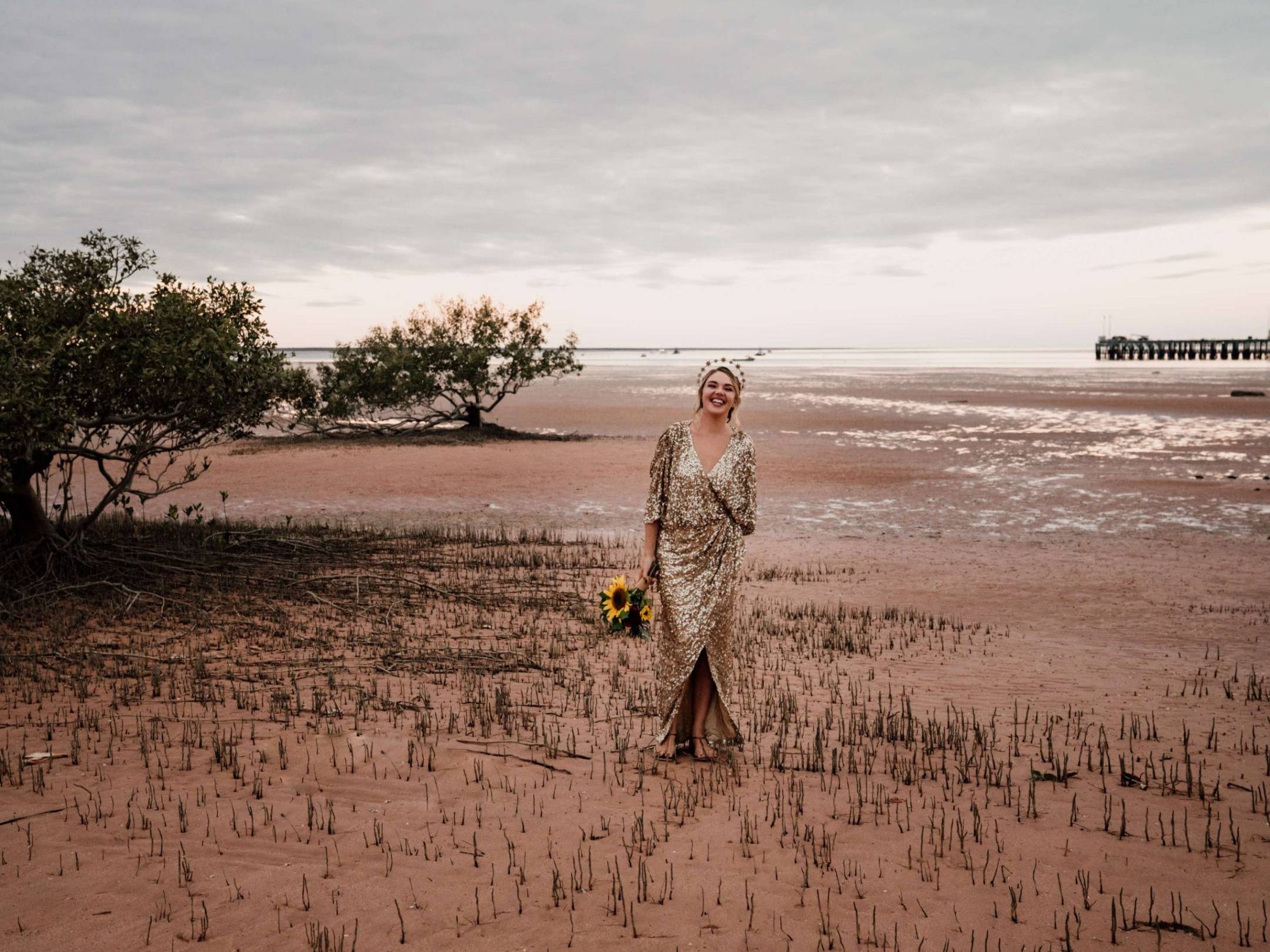 Woman in a gold dress with sunflowers on a beach, with a pier in the background.