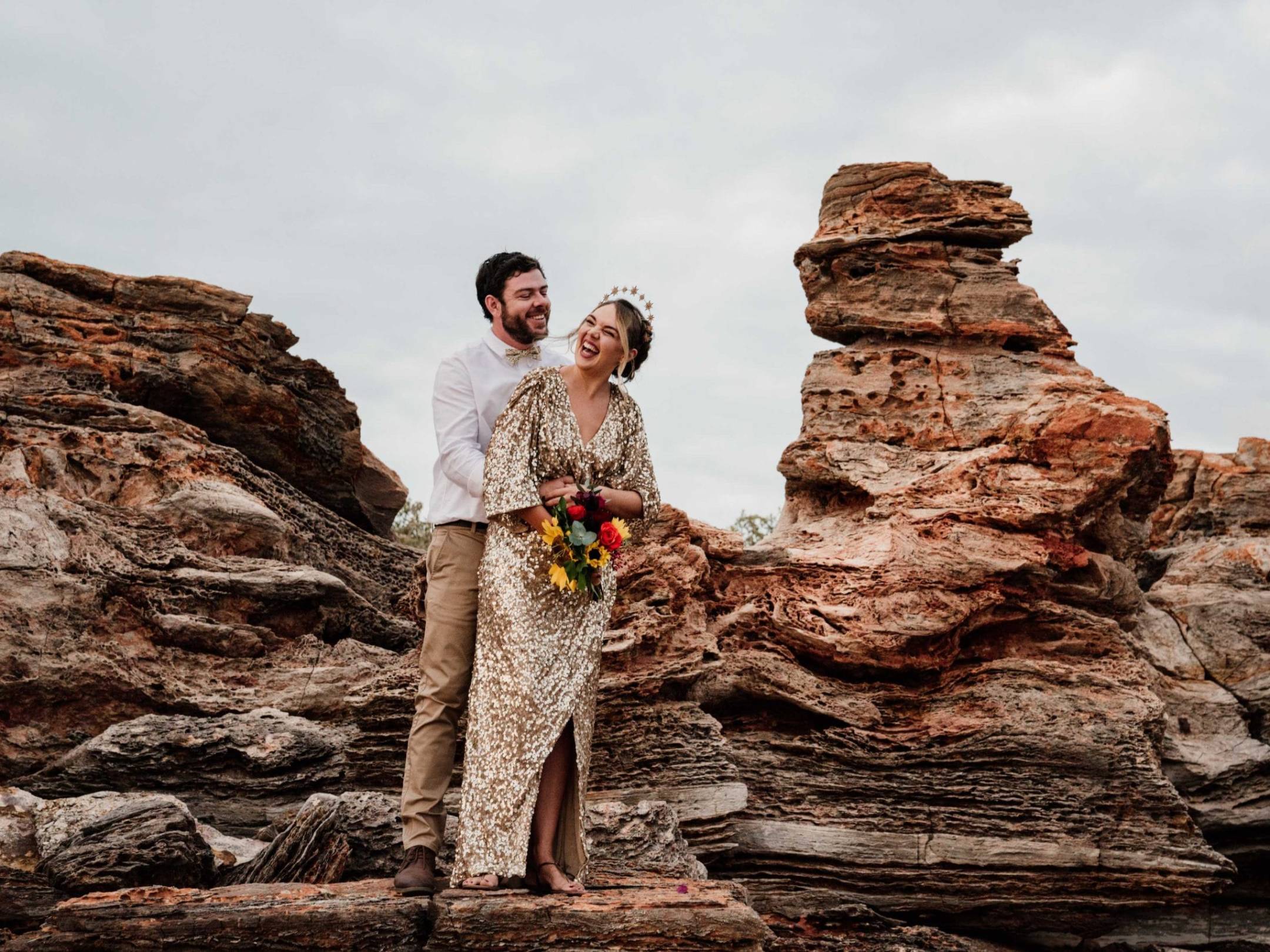 Couple smiling on rocky terrain, woman holding bouquet in sequined dress, man in white shirt.