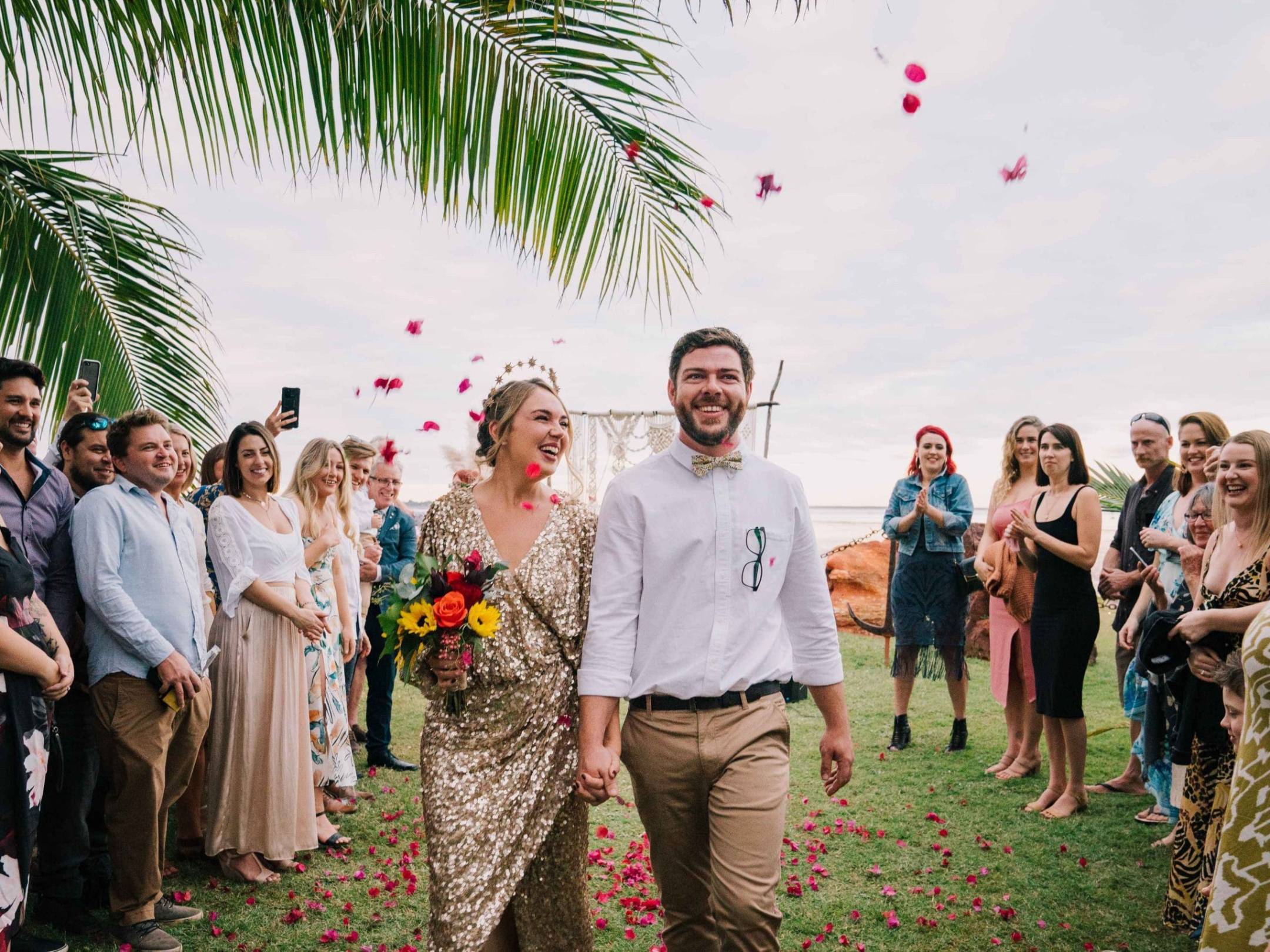 A couple walks down an aisle outdoors, surrounded by smiling guests and petals falling.