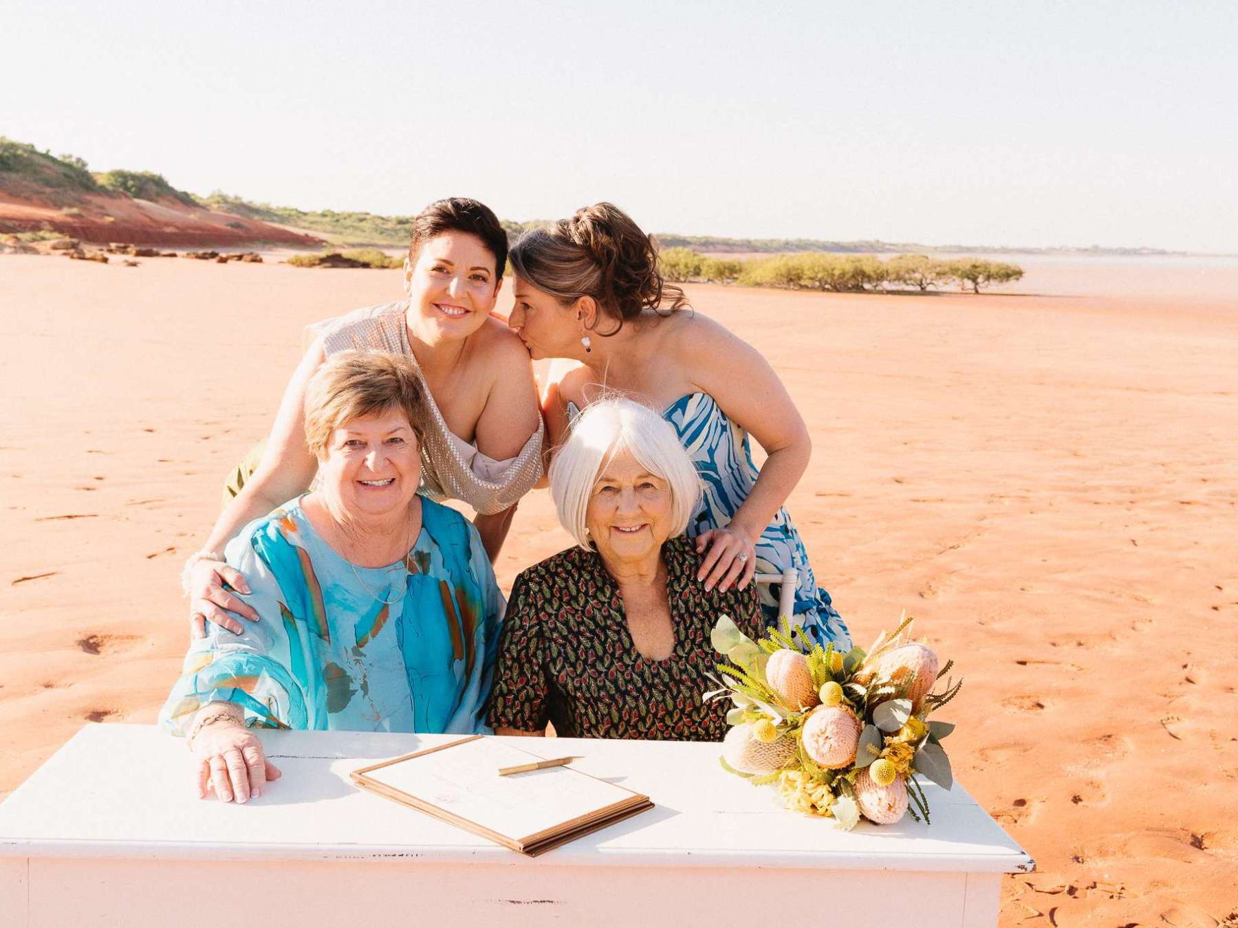 Four women posing on a sandy beach with a table and flowers.