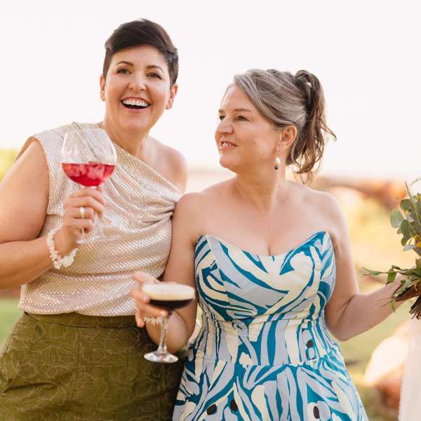 Two women smiling, holding drinks. One holds flowers. They are outdoors in a garden setting.