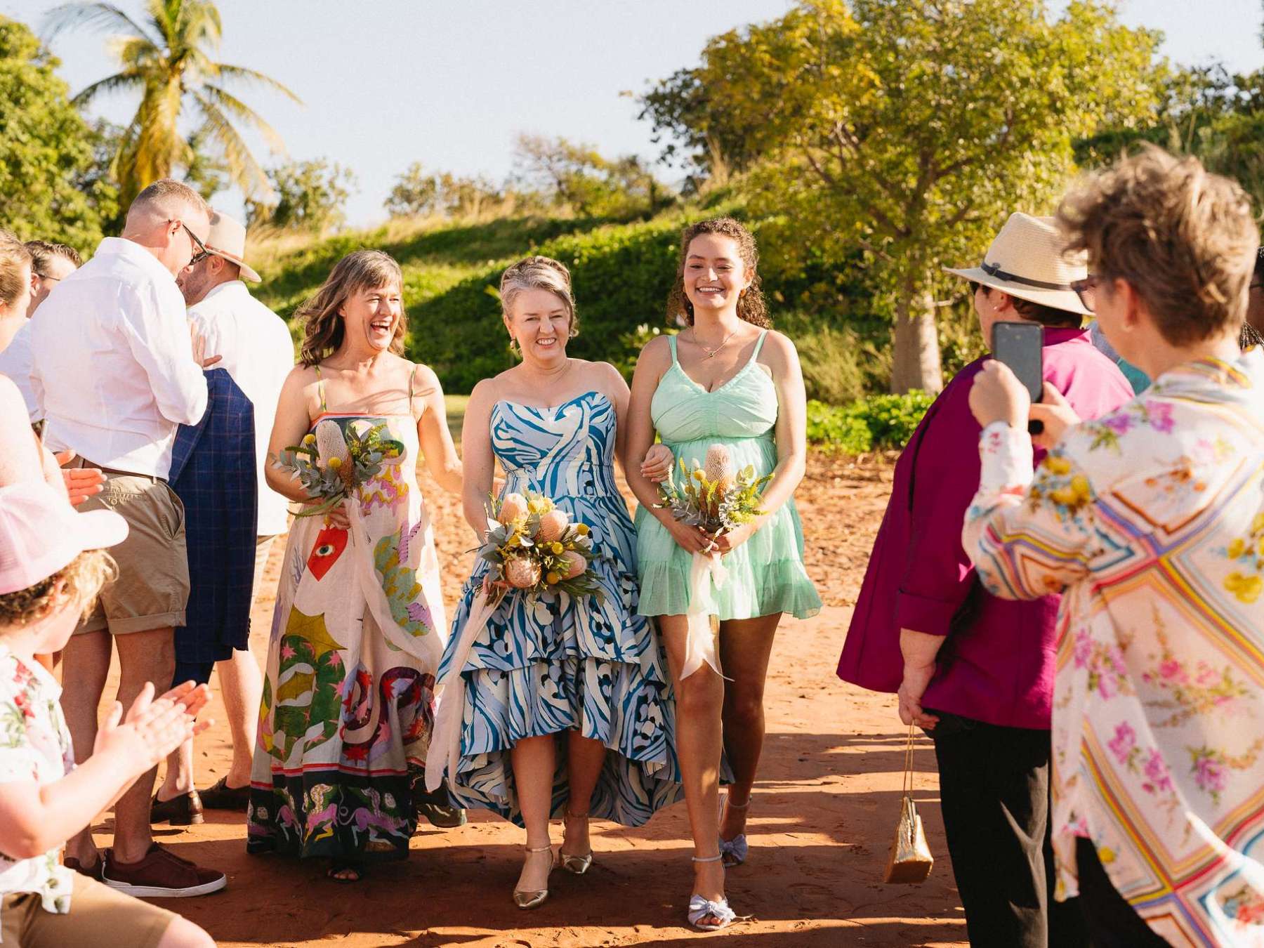 Three women in colorful dresses walk down a sandy path surrounded by applauding people.