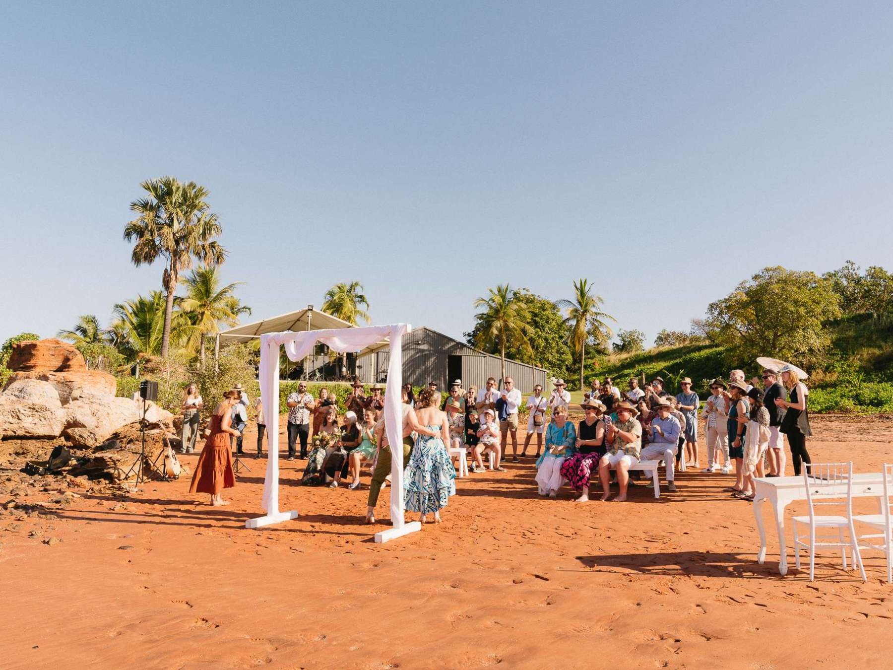 Outdoor wedding ceremony on sandy beach with guests and white arch.
