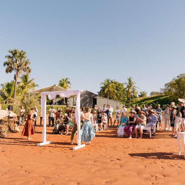 Outdoor wedding ceremony on sandy beach with guests and white arch.