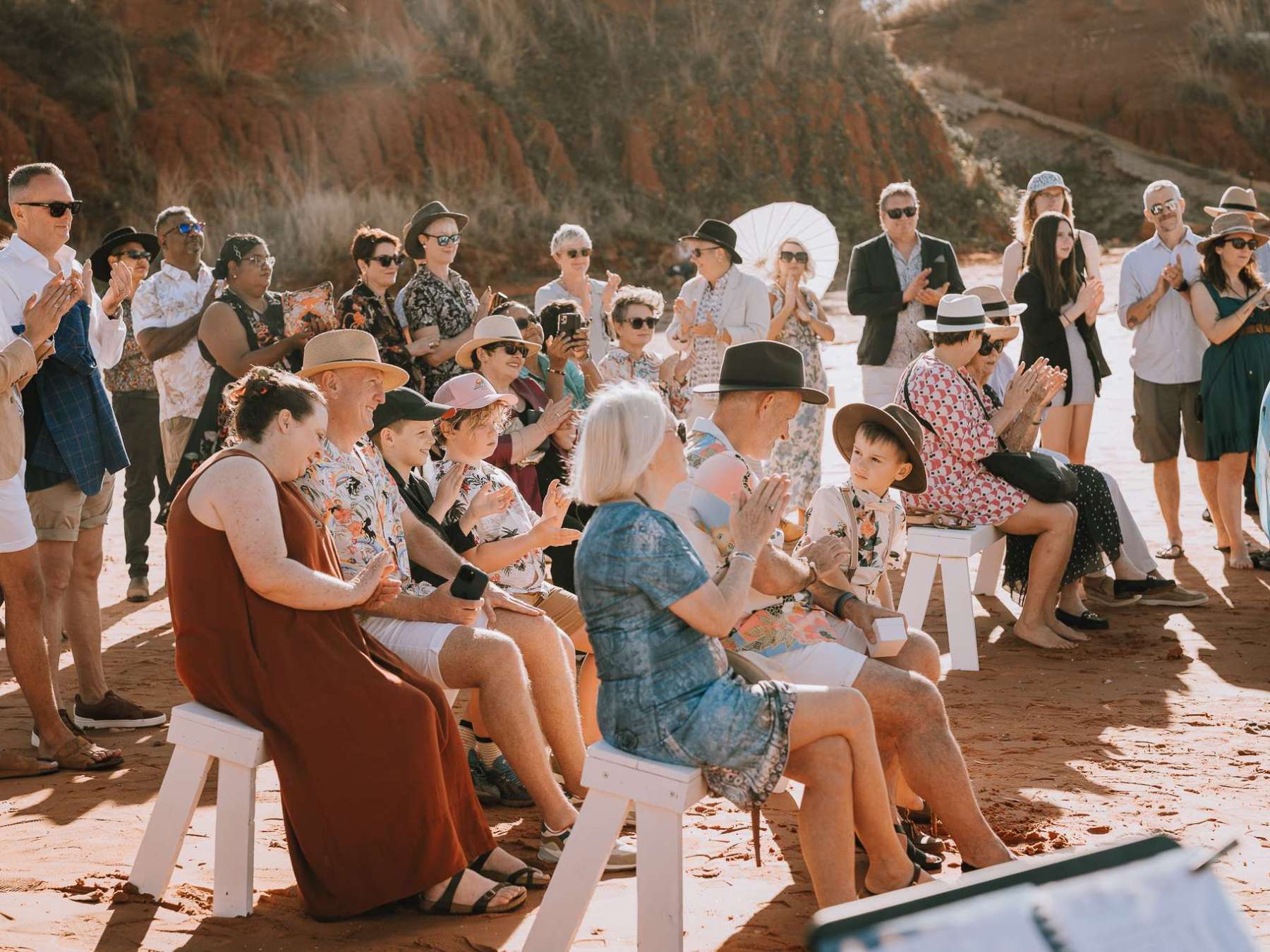 A group of people sitting and standing outdoors, clapping and wearing casual summer attire.
