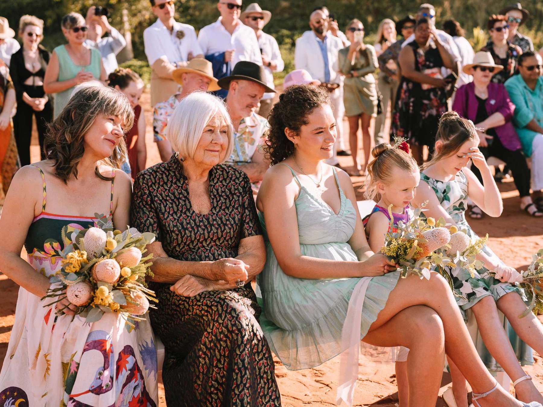 A group of women and children seated outdoors, holding bouquets at an event with people standing in the background.