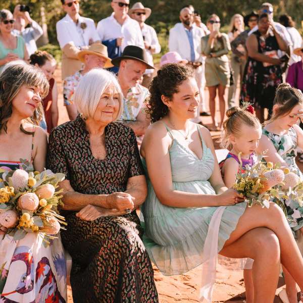 A group of women and children seated outdoors, holding bouquets at an event with people standing in the background.