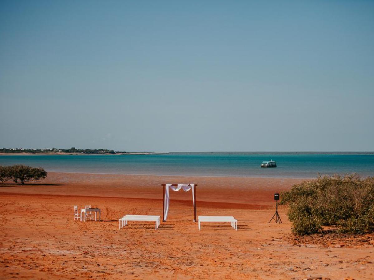 Sunlit beach wedding setup with a small canopy, chairs, and tables, overlooking a calm blue sea.