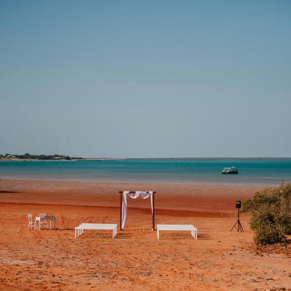 Sunlit beach wedding setup with a small canopy, chairs, and tables, overlooking a calm blue sea.