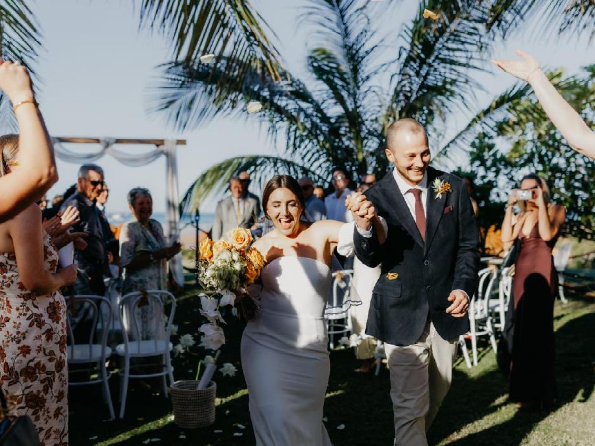 A couple walks hand in hand at a sunny outdoor wedding with cheering guests and palm trees.