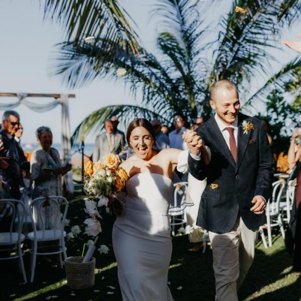 A couple walks hand in hand at a sunny outdoor wedding with cheering guests and palm trees.