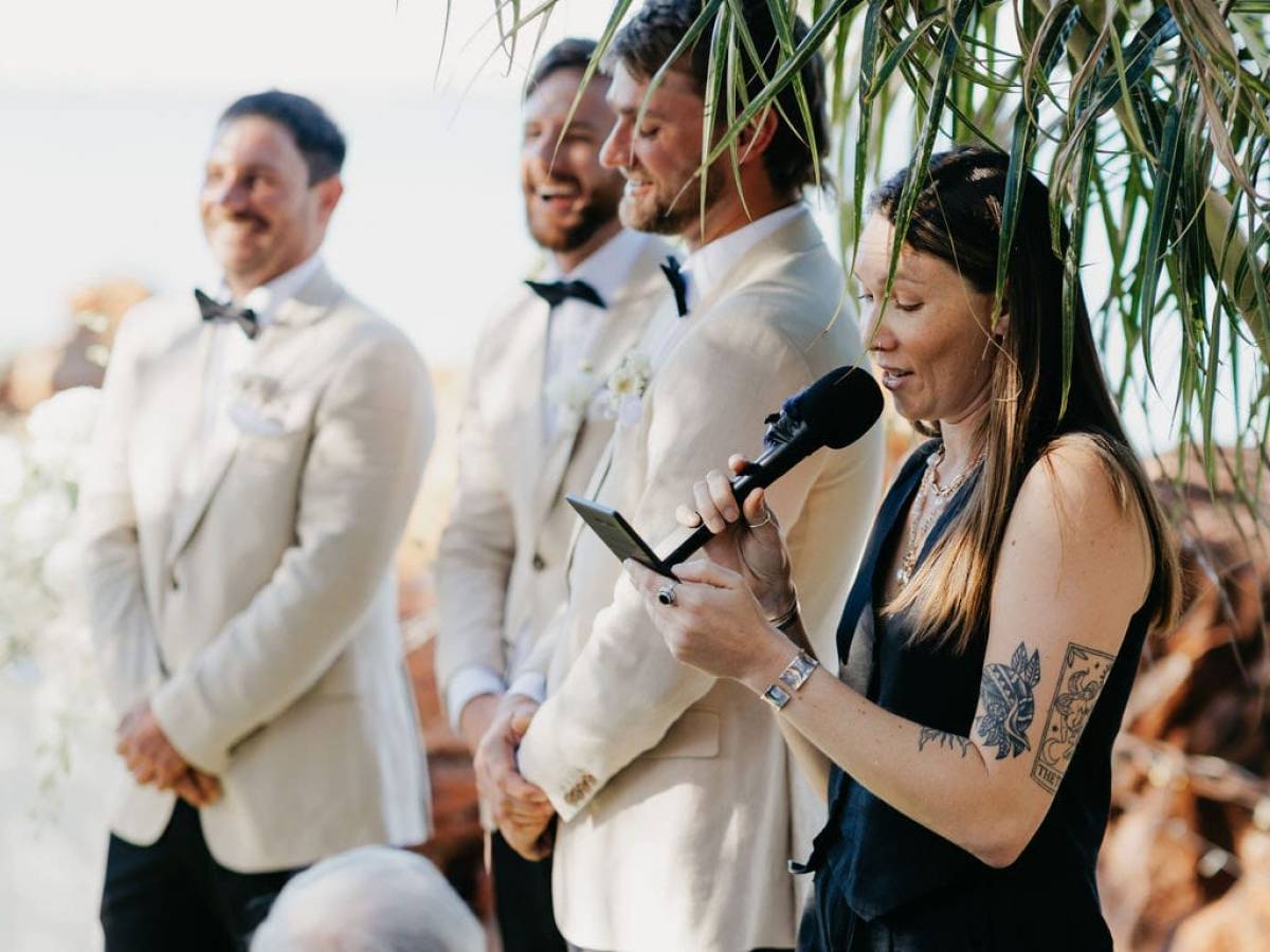 Person speaking into a microphone next to three people in suits under palm leaves at an outdoor event.