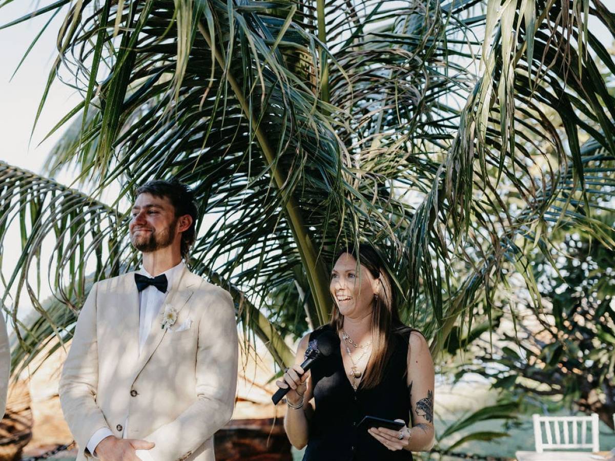 Person in suit and another with a microphone under a palm tree, outdoors.
