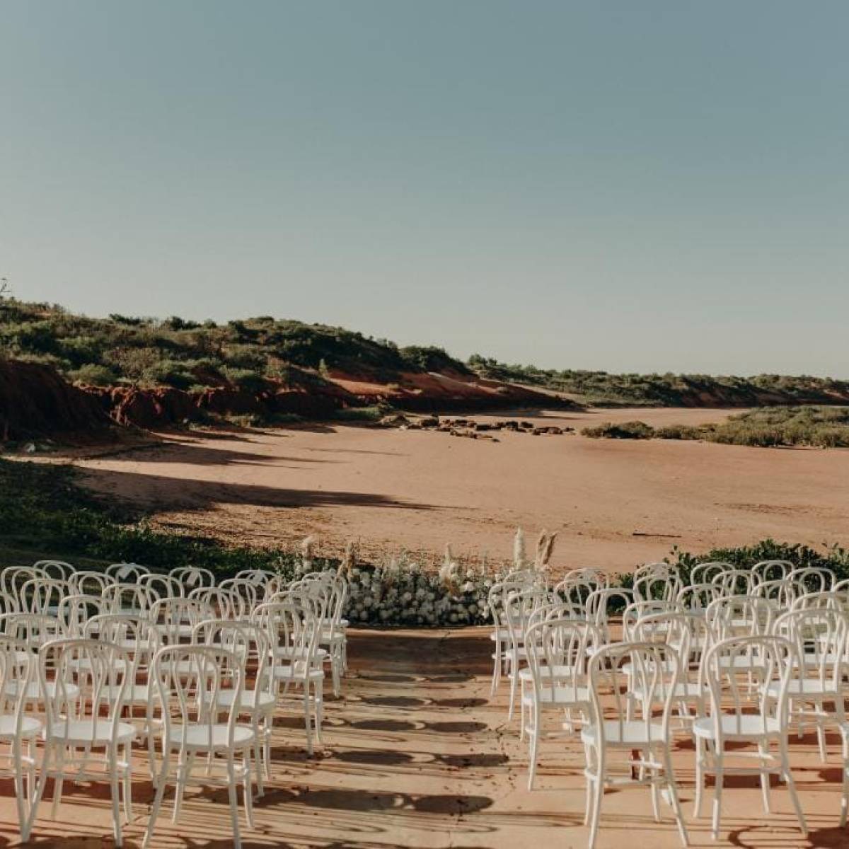 Outdoor wedding setup with white chairs facing an empty sandy landscape and clear sky.