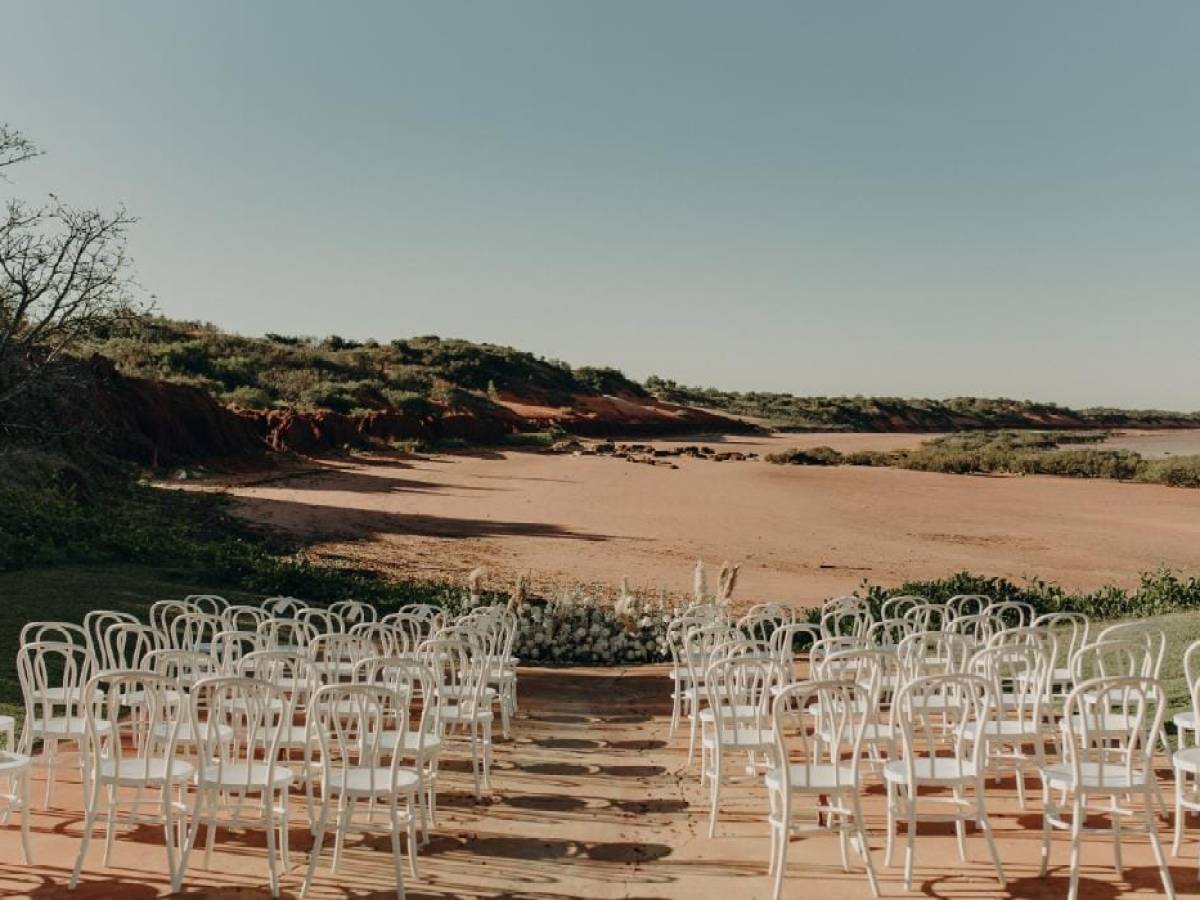 Outdoor wedding setup with white chairs facing an empty sandy landscape and clear sky.