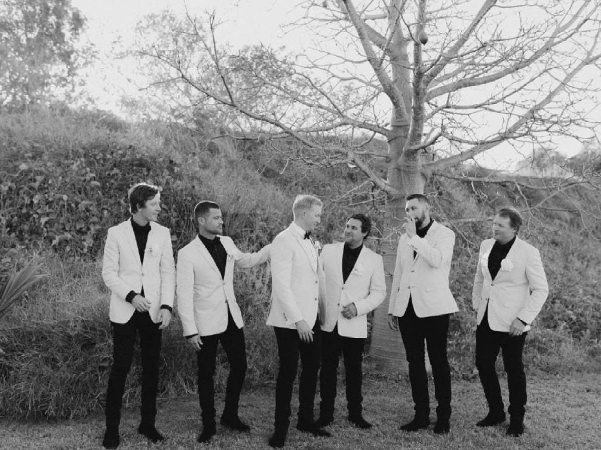 Six men in suits standing in a field, laughing near a leafless tree.