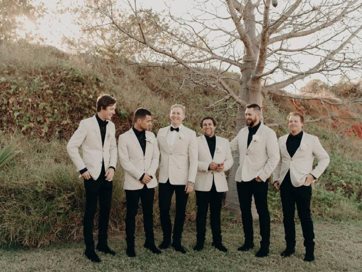 Six men in beige suits and black shirts stand smiling outdoors near a tree.