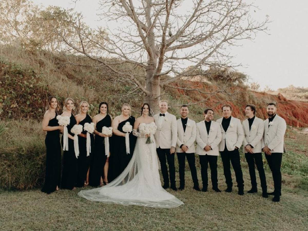Wedding party stands outside; bridesmaids in black, groomsmen in white jackets, with a bride and groom in the center.