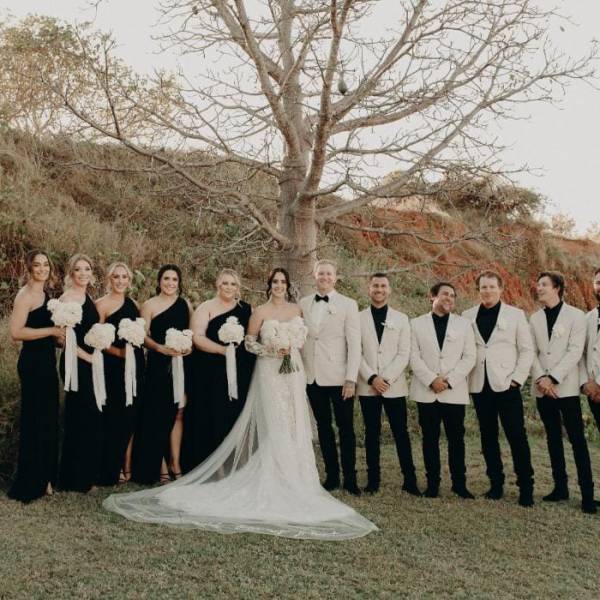 Wedding party stands outside; bridesmaids in black, groomsmen in white jackets, with a bride and groom in the center.
