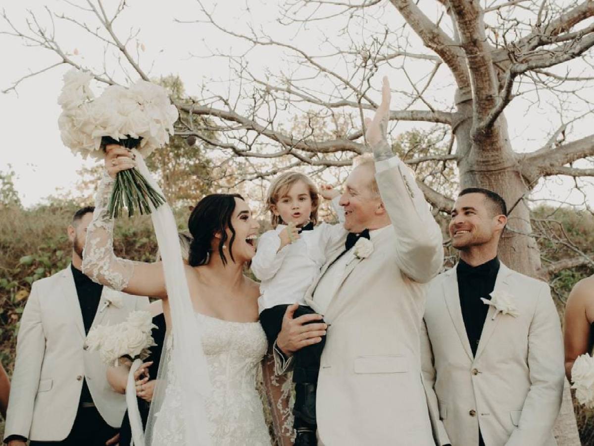 Bride and groom celebrating with a child and guests under a tree.