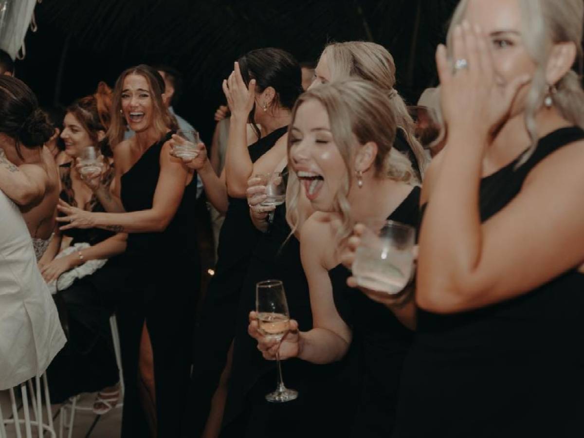 Group of women in black dresses laughing and holding drinks at an event.