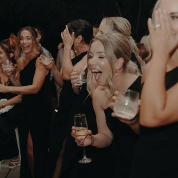 Group of women in black dresses laughing and holding drinks at an event.