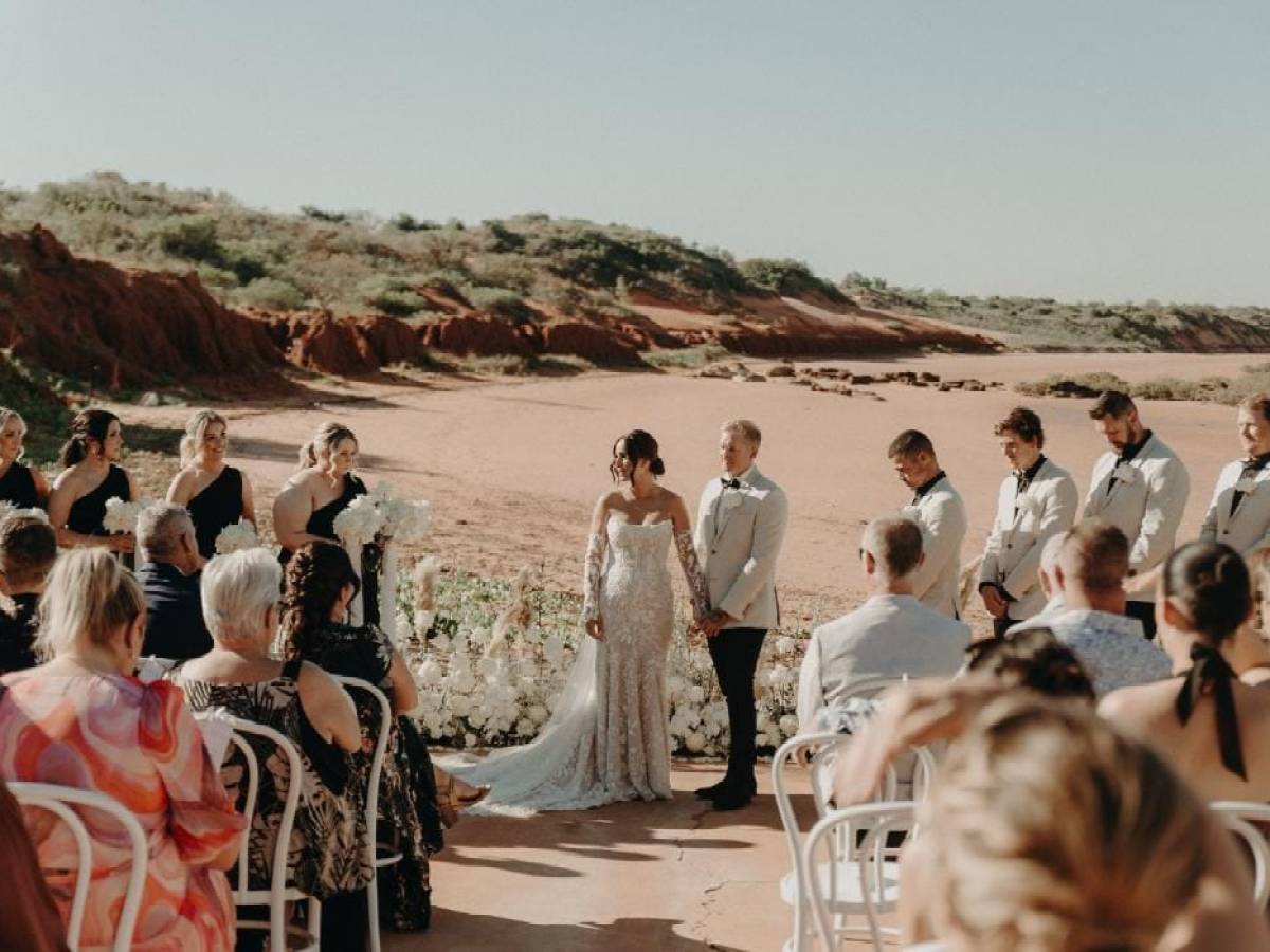 Outdoor wedding ceremony with a couple holding hands, bridesmaids in black, groomsmen in light suits, guests seated.