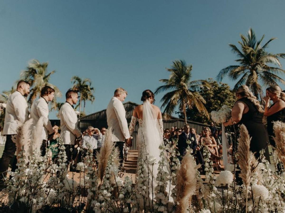 Outdoor wedding ceremony with bride, groom, and guests surrounded by palm trees.