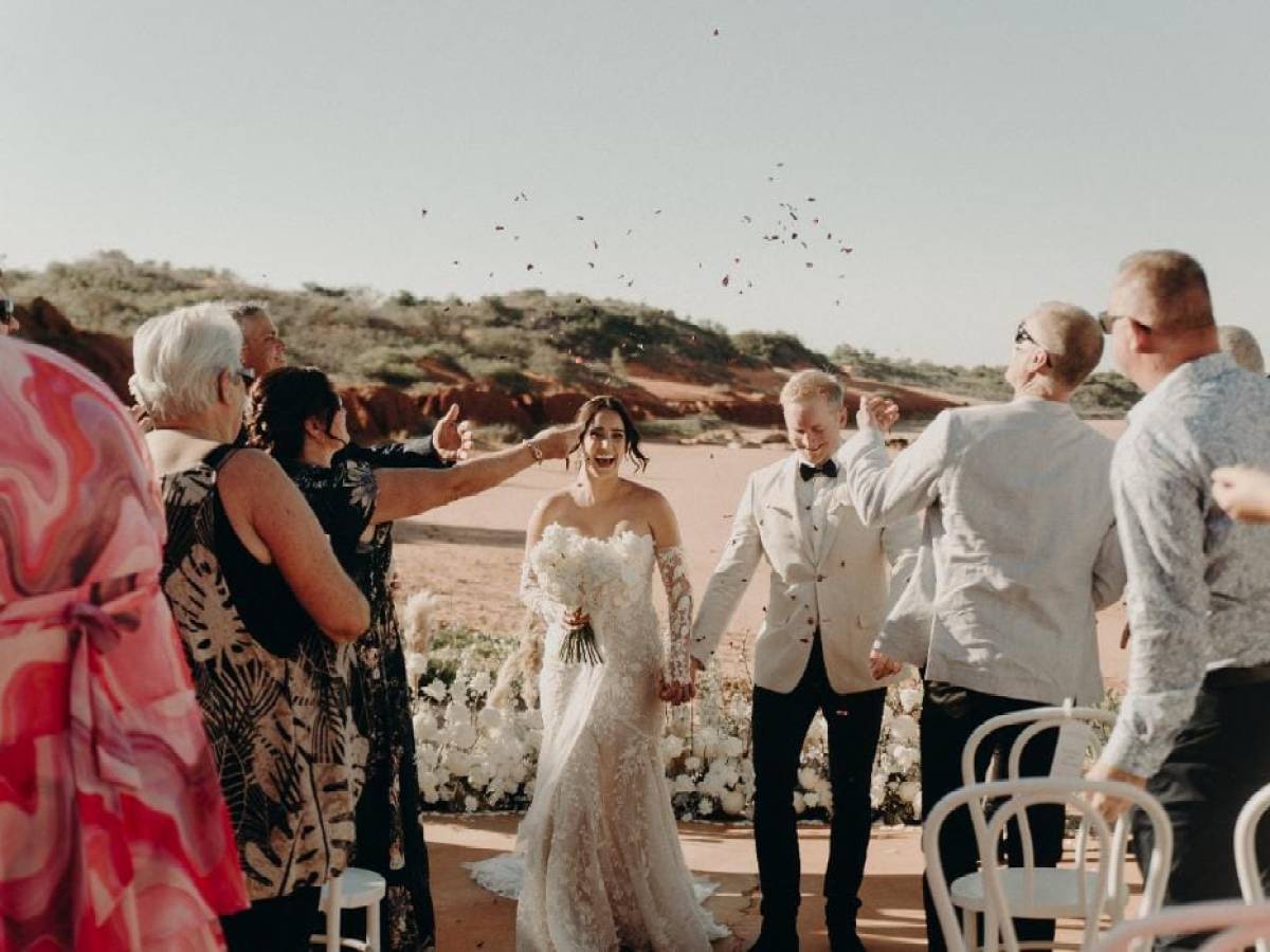 Bride and groom walk down an outdoor aisle with guests cheering and throwing confetti.