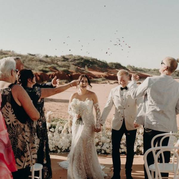 Bride and groom walk down an outdoor aisle with guests cheering and throwing confetti.